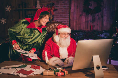 Photo portrait of santa claus and elf looking at computer screen holding bunch of lettersの写真素材