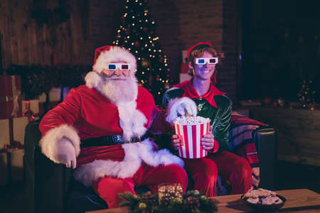 Photo portrait of santa claus and elf eating popcorn watching movie on sofa in 3d glassesの写真素材
