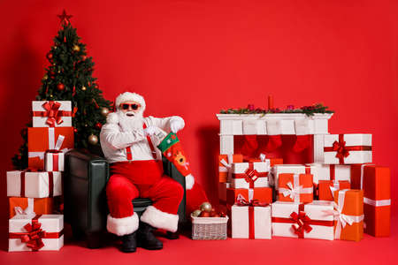 Portrait of his he attractive fat overweight cheerful glad Santa sitting in armchair living-room packing us cash gift in ugly sock isolated bright vivid shine vibrant red color backgroundの写真素材