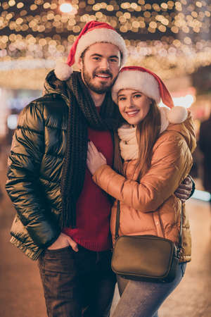 Affectionate jolly couple hugging on x-mas fair under evening outside tradition lights wear winter coats santa claus headwearの写真素材