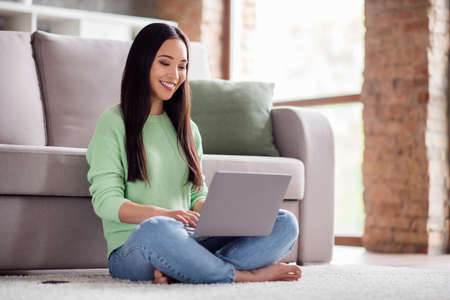 Full length profile side photo of positive korean girl sit floor carpet legs crossed folded barefoot work remote laptop have training web meeting in house indoorsの写真素材