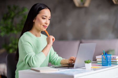 Photo of focused asian girl sit cozy desk work study remote prepare report hold pen copybook look laptop screen in house indoorsの写真素材
