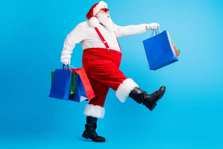 Full body profile side photo of crazy white grey hair bearded fat santa claus go shopping hold x-mas christmas bags wear red suspenders overalls isolated over blue color backgroundの写真素材