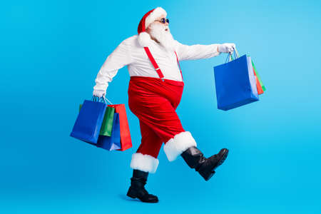 Full length photo of modern white grey hair bearded santa claus go shopping hold x-mas christmas bags wear headwear suspenders overalls boots isolated over blue color backgroundの写真素材