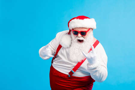 Crazy stylish santa claus with big stomach beard show horns symbol x-mas christmas jolly fun wear style trendy suspenders overalls headwear isolated blue color backgroundの写真素材