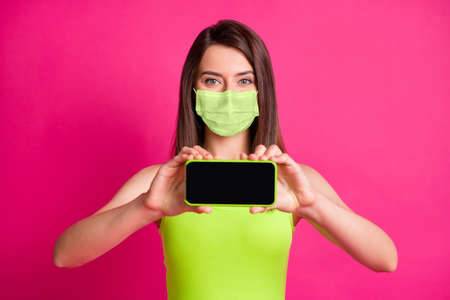 Photo of charming young lady hold phone show screen empty space wear green face mask isolated bright pink color backgroundの写真素材