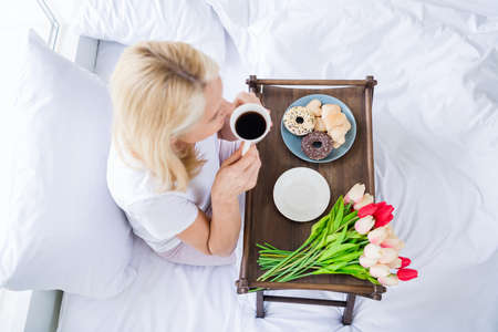 High angle view photo of woman sit bed linen drink fresh coffee tray tulips cookies bedroom indoors
