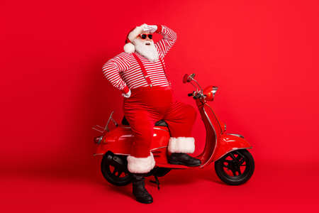 Full length photo of grandfather grey beard stand near retro scooter look far away wear santa claus x-mas costume suspenders sunglass striped shirt boots cap isolated red color backgroundの写真素材