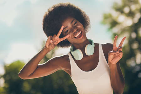 Portrait of positive cheerful dark skin girl make v-sign near face in summer woods park wear white tank-topの写真素材