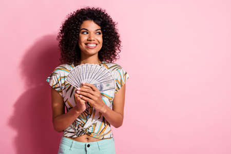 Portrait photo of black skinned curly curious girl keeping showing money looking at blank space isolated on pastel pink color backgroundの写真素材