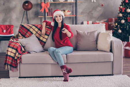 Photo of girl sit couch hold remote control point you look camera wear santa cap red pullover jeans socks in decorated living room indoorsの写真素材
