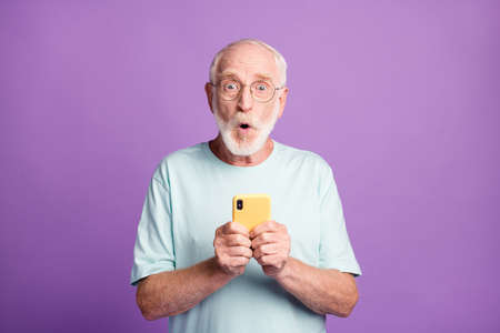 Photo portrait of shocked old man holding phone in two hands isolated on vivid violet colored backgroundの写真素材