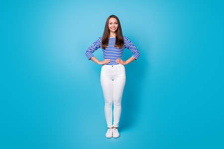 Full length photo of good-looking healthy young girl fit figure hands hips sides guide cruise liner meet passengers wear striped shirt white trousers sneakers isolated blue color backgroundの写真素材