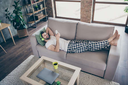 Top above high angle view full body photo of positive guy lie divan use smartphone listen radio wireless headset wear checkered plaid pants trousers white t-shirt in house indoorsの写真素材