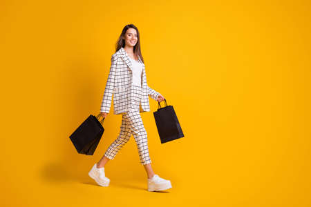 Full length profile photo of cheerful girl walk shopping center hold many bags wear checkered cloth white sneakers isolated over yellow color backgroundの写真素材