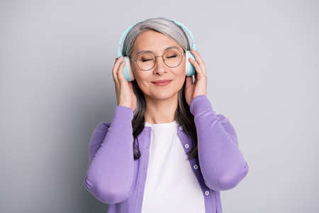 Photo portrait of elder woman listening to music enjoying touching wireless earphones isolated on grey color backgroundの写真素材
