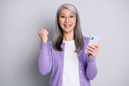 Photo portrait of cheerful woman celebrating victory holding mobile phone one fist up isolated on grey colored backgroundの写真素材