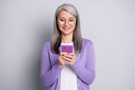 Portrait photo of smart grandmother using smartphone texting browsing wearing eyeglasses smiling isolated on grey color backgroundの写真素材