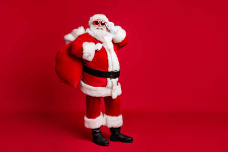 Full length photo of retired old man grey beard hold glasses bear sack presents good children wear santa costume gloves coat belt sunglass headwear black boots isolated red color backgroundの写真素材