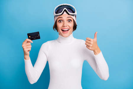 Photo of brunette lady wear white turtleneck mask goggles hold credit card show thumb up isolated on blue color backgroundの写真素材