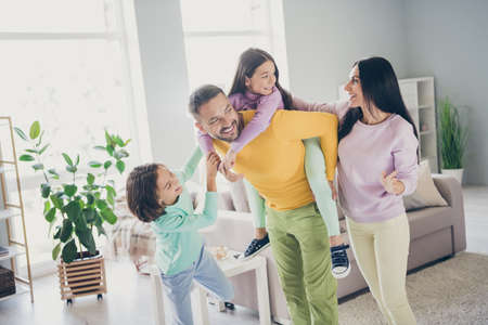 Photo of full family four members daddy hold piggy back daughter enjoy weekend wear colorful jumper trousers in living room indoorsの写真素材