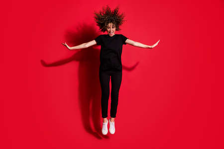 Photo portrait full body view of happy girl jumping up with hands to both sides isolated on vivid red colored backgroundの写真素材