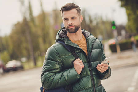 Photo of good mood confident young man wear green windbreaker schoolbag holding modern device outside city streetの写真素材