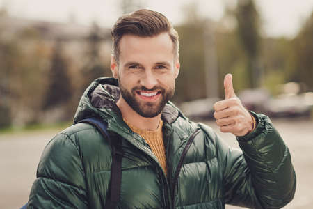 Photo of toothy beaming young guy dressed green coat rucksack walking showing thumb up one hand outdoors urban city streetの写真素材