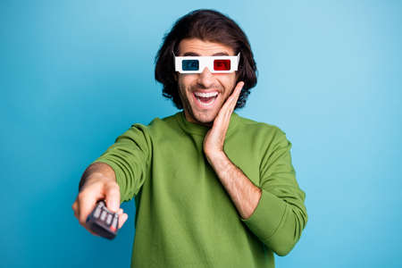 Photo of amazed funky man hold remote control palm cheek wear 3d glasses green pullover isolated blue color backgroundの写真素材