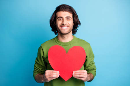 Photo of cheerful young man beaming smile show paper heart wear green pullover isolated blue color backgroundの写真素材
