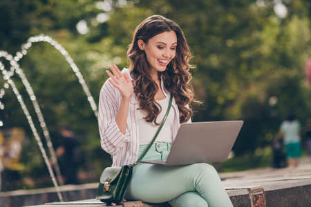 Photo of nice cheerful brunette lady sit near fountain talking laptop wear pastel cloth shoulder bag in park outdoorsの写真素材