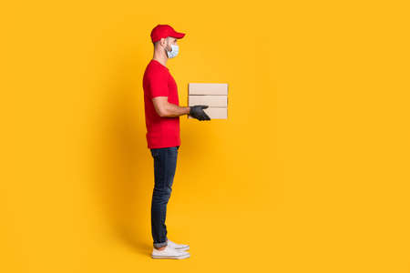 Profile photo of courier man hold carton boxes wear mask red t-shirt hat jeans footwear isolated yellow color backgroundの写真素材