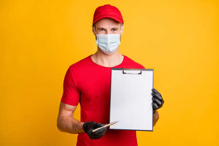 Photo of young delivery guy hold clipboard paper empty space pen wear mask red t-shirt hat isolated...