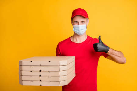 Photo of young courier man hold pizza boxes raise thumb up wear mask red t-shirt cap isolated yellow color backgroundの写真素材