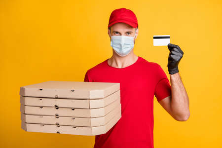 Photo of courier man hold pizza boxes debit card wear red t-shirt headwear gloves isolated yellow color backgroundの写真素材