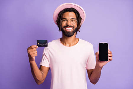 Photo portrait of man in stylish hat headwear cap holding credit card phone with copyspace on display isolated on vivid violet colored backgroundの写真素材