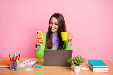Positive girl high school student sit table use smartphone hold mug wear green shirt isolated pink color backgroundの写真素材