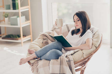 Full length photo of concentrated girl sit on wicker chair read book hold coffee cup in modern house...