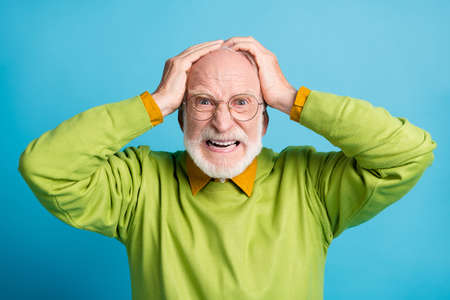 Photo of aggressive retired old man hands head mad look wear glasses green sweater isolated blue...