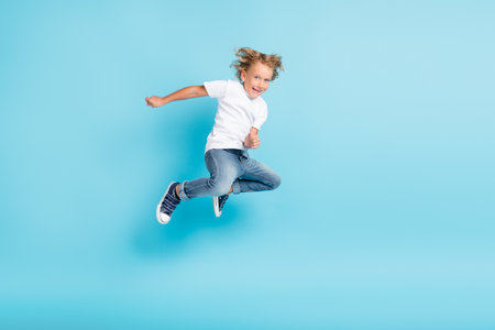 Little kid jump motion funny grimace hands wear white shirt jeans sneakers isolated blue color backgroundの写真素材