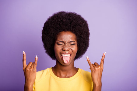Portrait of crazy dark skin girl make horned symbol show tongue wear yellow sweater isolated over purple color backgroundの写真素材