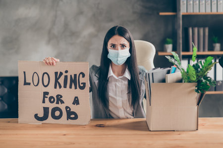I need work. Frustrated stressed girl ceo marketer banker sit table desk lose job company covid economics crisis show cardboard text box wear medical mask blazer jacket in workplaceの写真素材