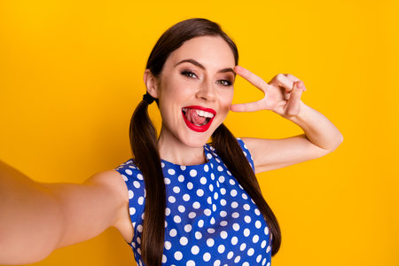 Self-portrait of her she nice-looking attractive pretty lovely glamorous funky cool cheerful girl showing v-sign grimacing having fun isolated on bright vivid shine vibrant yellow color backgroundの写真素材