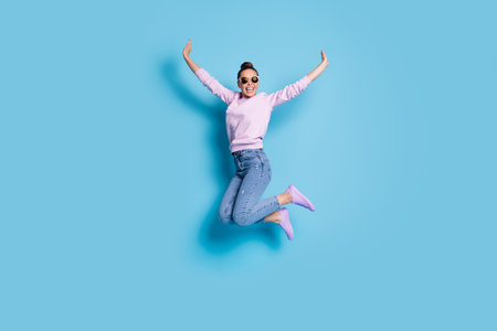 Full length photo of cheerful candid energetic girl jump enjoy weekend raise hands wear good look pullover isolated over blue color backgroundの写真素材