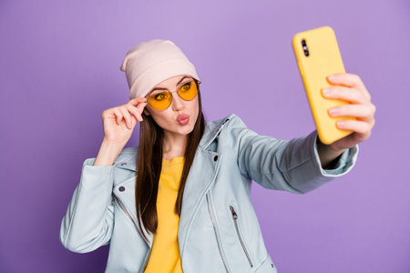 Photo of funny pretty youngster lady hold telephone make selfies popular blogger sending air kisses wear sun specs casual hat jacket yellow pullover isolated purple color backgroundの写真素材