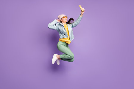 Full length photo funny foolish girl have autumn free time jump make selfie smartphone show tongue out wear leather jacket sunglass green pants trousers isolated violet color backgroundの写真素材