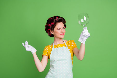 Photo positive house wife girl wash wine glass look enjoy perfect neat tableware polishing wear yellow dotted style dress hair rollers isolated over green color backgroundの写真素材