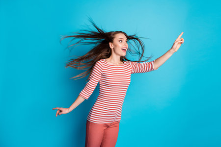 Photo of astonished girl see incredible advertisement promo point index finger copyspace her hairdo wind air throw blow wear stylish trendy outfit isolated over blue color backgroundの写真素材