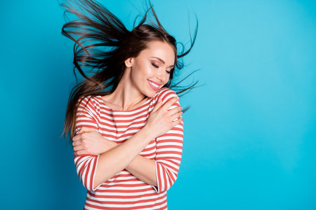 Photo of satisfied candid girl hug herself enjoy cuddle love her hair fly wind blow isolated over blue color background wear look good mood shirtの写真素材