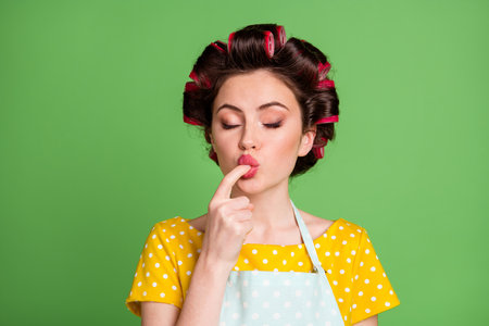 Photo of charming pretty student girl roller hairstyle lips lick finger tasting cream closed eyes enjoying perfect taste wear dotted apron shirt isolated green color backgroundの写真素材